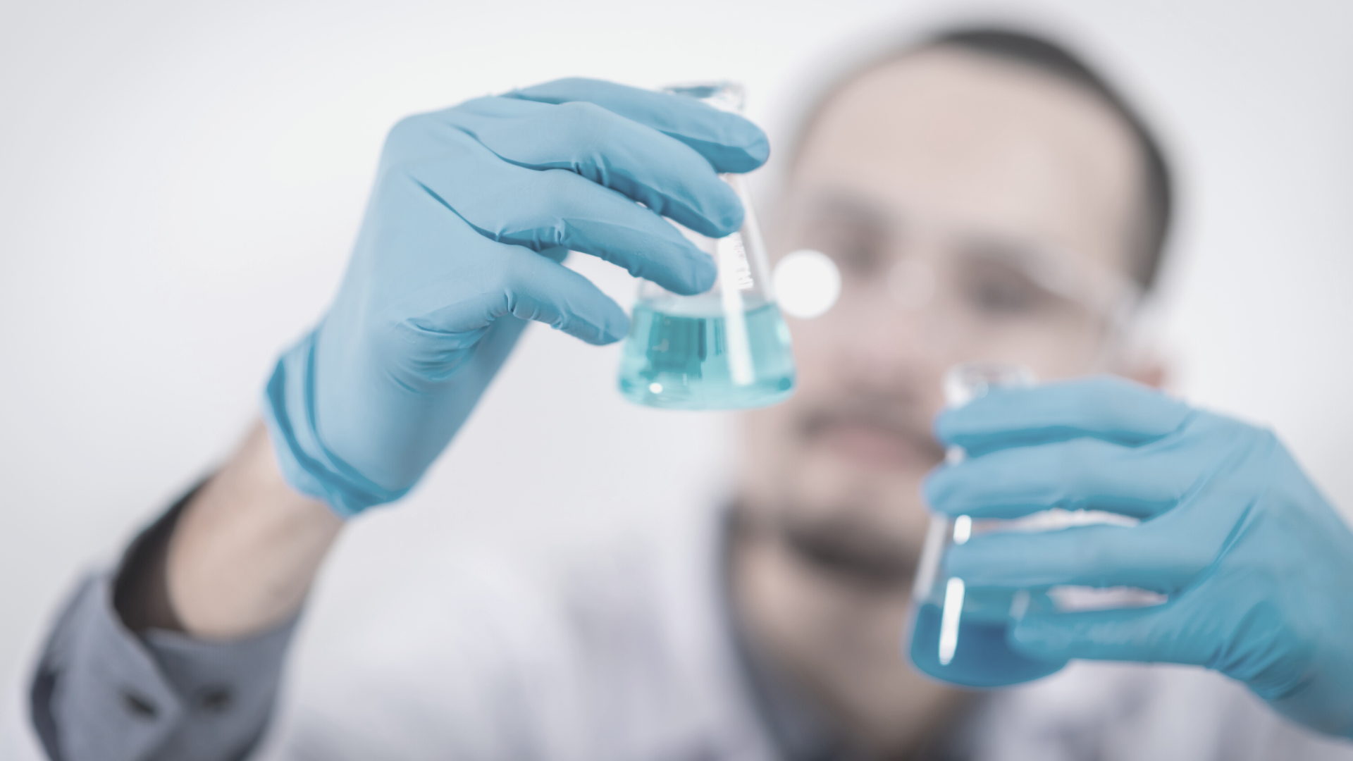 man wearing safety goggles and blue gloves holding two test tubes containing blue liquid